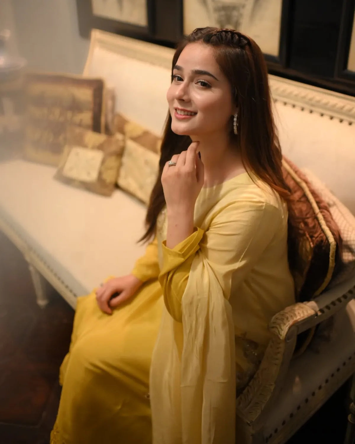 A woman wearing a yellow traditional outfit sits on a cushioned bench in an indoor setting with warm lighting.