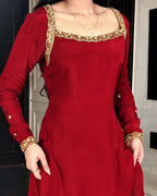 A woman wearing a long-sleeved red dress with ornate gold embroidery around the square neckline and cuffs, shown from the shoulders to mid-thigh in an indoor setting.
