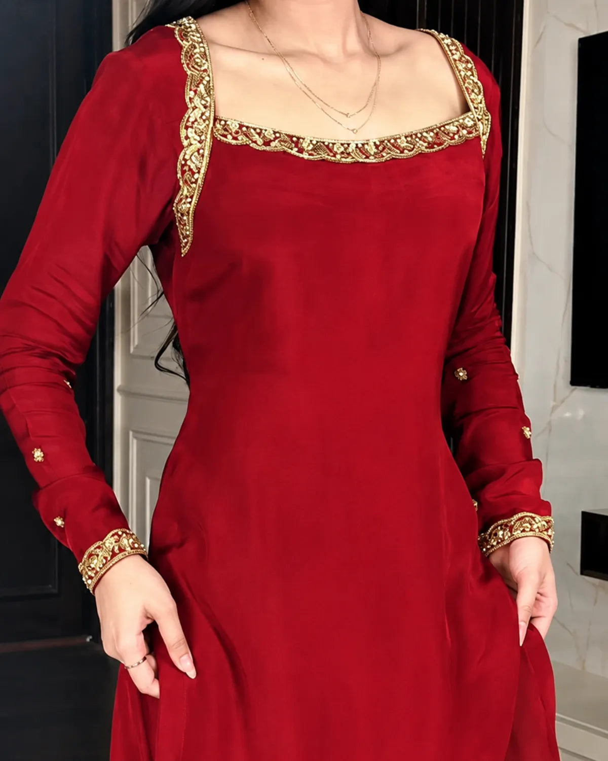 A woman wearing a long-sleeved red dress with ornate gold embroidery around the square neckline and cuffs, shown from the shoulders to mid-thigh in an indoor setting.