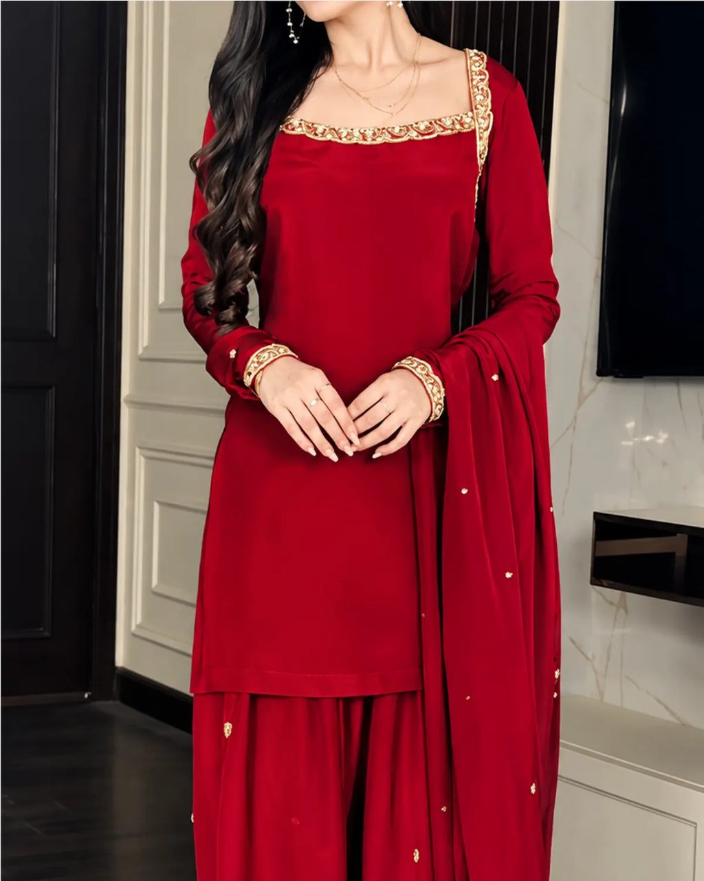 A woman wearing a deep red three-piece ethnic suit with gold embroidery on the neckline, sleeve trim, and dupatta, standing in an indoor setting.