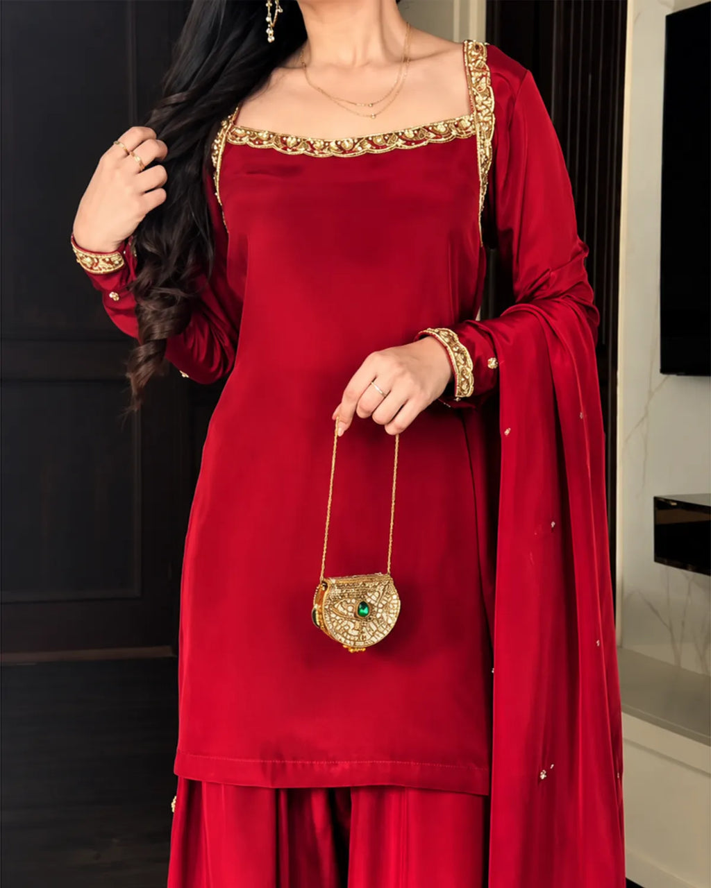 A woman wearing a deep red traditional outfit with gold embroidered trim, holding a small gold purse with a green stone accent.