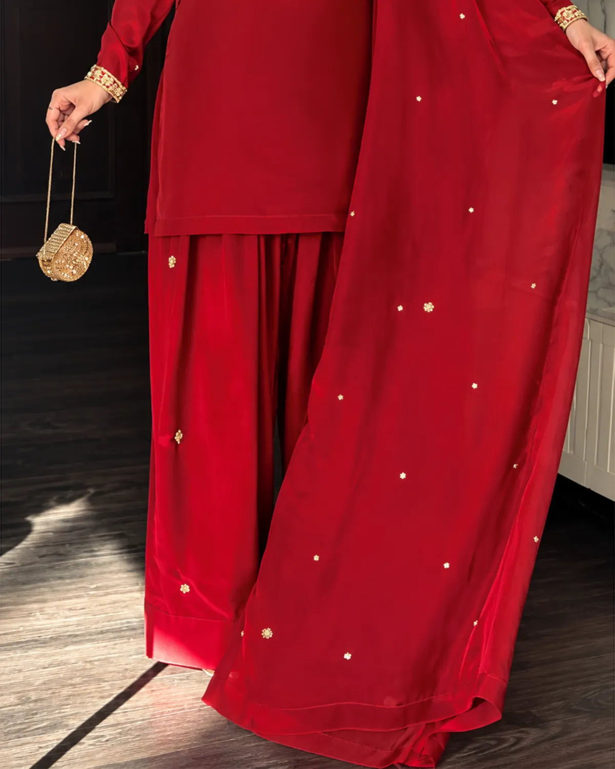 A close-up of a red traditional outfit with wide-leg pants and a matching draped fabric, both decorated with small gold embroidered details. A hand holding a small gold purse is visible on the left, standing on a dark wood floor.