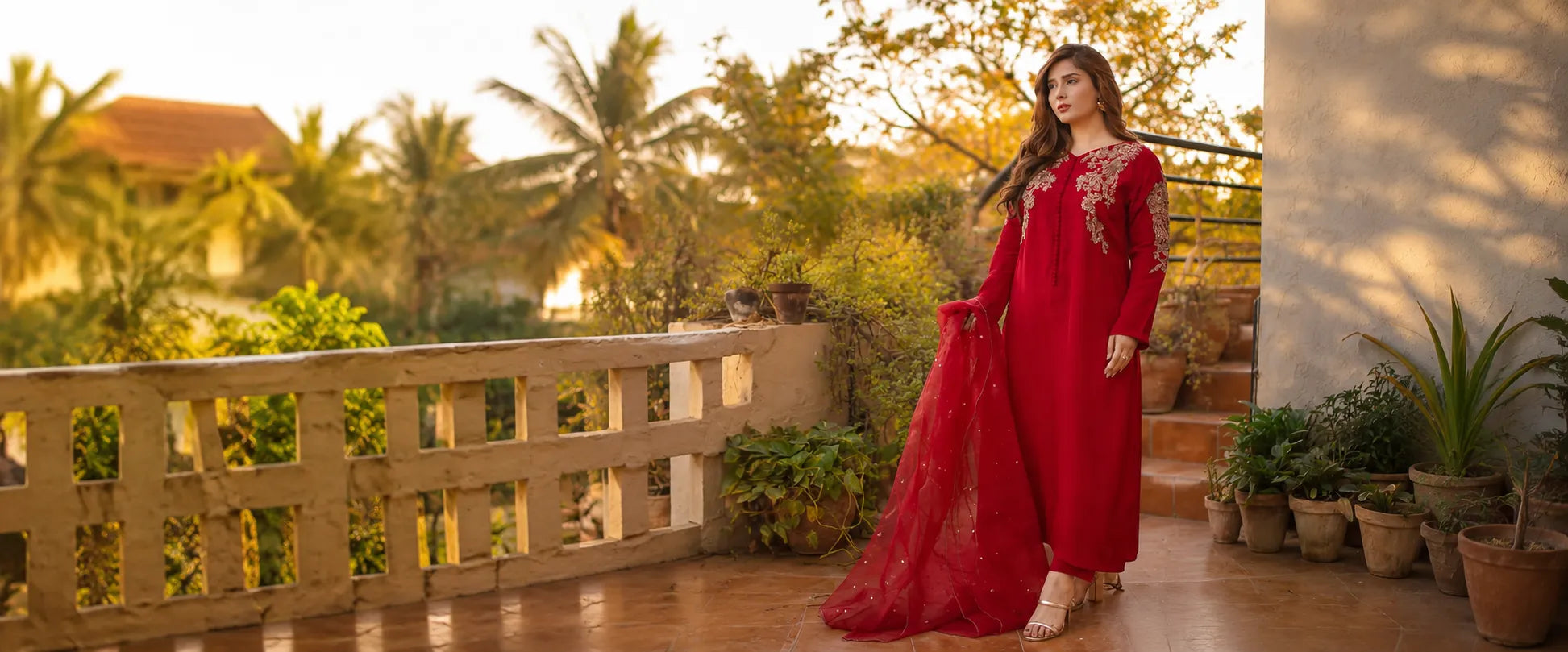 A woman wearing a long red embroidered salwar kameez suit stands on a sunlit balcony with plants and a house in the background.